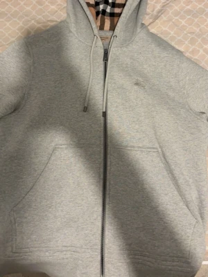 Grå kofta - Grå zip-hoodie med mönstrad luva Säljer en stilren och kraftig hoodie i ljusgrå melerad färg. Tröjan har en klassisk passform med hel dragkedja framtill och två rymliga fickor. 