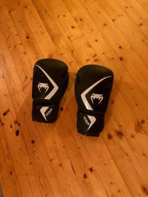 Svarta boxningshandskar från Venum - Säljer ett par svarta boxningshandskar från Venum med vita detaljer och logga på ovansidan. Handskarna har kardborreknäppning vid handleden för enkel justering och bra stöd. Perfekta för boxning eller annan kampsportsträning.Mycket bra skick knappast använda.