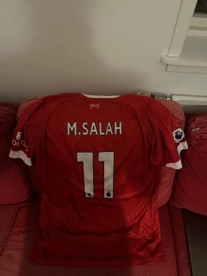 Liverpool matchtröja M. Salah 11 Nike ÄKTA - WSäljer en röd Liverpool matchtröja från Nike med M. Salah och nummer 11 på ryggen. Tröjan har vita detaljer vid krage och ärmslut samt Premier League-logga på ärmen. Perfekt för fotbollsfans som vill stötta sitt lag.