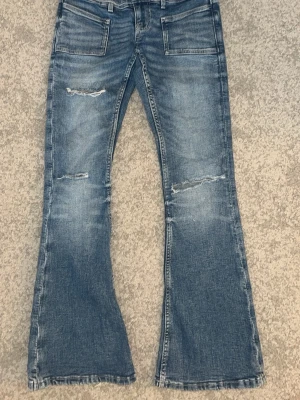 Blå utsvängda jeans med slitningar - Säljer ett par blå jeans med utsvängda ben och slitningar vid knäna. Jeansen har stora framfickor och klassiska bakfickor. Materialet är denim i bomull och färgen är tvättad blå med ljusare partier. 