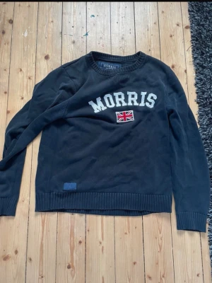 Mörkblå stickad tröja Morris - Snygg mörkblå stickad tröja från Morris med vit text på bröstet och en liten brittisk flagga. Tröjan har rund halsringning, ribbade muddar och nederkant samt en liten blå patch nertill.  Finns skador på tröjan se bild. 