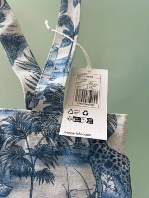 Blåvit shoppingväska från Stronger - Snygg shoppingväska från Stronger i blåvitt tyg med tropiskt djungelmönster och djurmotiv. Väskan har två långa axelband och är tillverkad i tålig textil, perfekt för att bära med sig allt man behöver. Märkets logga syns framtill.