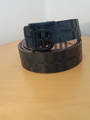 Svart rutigt Burberry bälte - Snyggt svart bälte från Burberry med klassiskt rutigt mönster och stor BB-logga i metall som spänne. Bältet är gjort i skinn och har präglade detaljer som ger en exklusiv känsla. Perfekt accessoar för att lyfta din outfit.fast pris 265kr