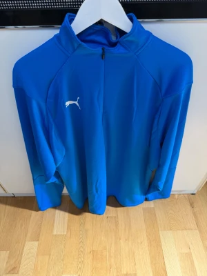 Blå träningsjacka från Puma - Snygg blå träningsjacka från Puma med halv dragkedja och hög krage. Tillverkad i ett lätt och stretchigt funktionsmaterial som passar perfekt till träning eller uppvärmning. Puma-logga i vitt på bröstet och lång ärm för extra komfort.