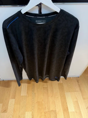 Svart långärmad träningströja SOC - Svart långärmad träningströja från SOC med diskret kamouflagemönster och en blå detalj på axeln. Tillverkad i ett lätt och ventilerande funktionsmaterial som passar perfekt för träning. Rund halsringning och enkel design.