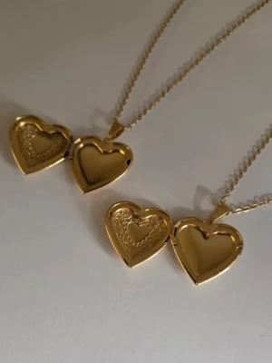 Två hjärthalsband med medaljong - Två matchande halsband i guldtonat rostfritt stål med hjärtformade medaljonger. Medaljongerna har graverade detaljer och kan öppnas för att lägga in små bilder eller minnen. Kedjan är tunn och elegant, perfekt för att bära varje dag. Kedjans längd är 50 cm och hänget ca 2,8 cm. 