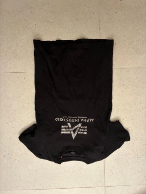 Svart Alpha Industries t-shirt - Snygg svart t-shirt från Alpha Industries med vit logga och text på bröstet. Klassisk rund hals och korta ärmar. Perfekt för dig som gillar streetwear och vill ha en enkel men stilren look.
