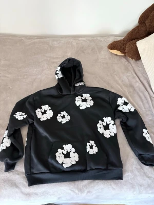 Svart hoodie from denim tears - Snygg svart hoodie med stora vita blommönster över hela tröjan. Hoodien har en klassisk huva och magficka framtill. Perfekt för dig som vill sticka ut med en unik och trendig stil. Materialet ser ut att vara mjukt och bekvämt, troligen polyester eller bomullsblandning.