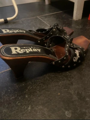Svarta träpumps från Replay - Säljer ett par svarta pumps från Replay med träklack och nitar längs remmarna. Skorna har öppen tå och Replay-logga på sidan. Snygg design med robust träbotten och dekorativa detaljer i metall.