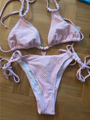 Ljusrosa bikini med pärldetaljer - Supersöt ljusrosa bikini med struktur och pärldetaljer i vitt och rosa på både överdel och underdel. Bikinin har knyt i sidorna och runt nacken för perfekt passform. Perfekt för sommaren och strandhäng! Aldrig använd. 