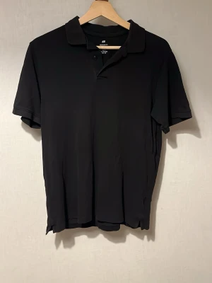 Svart pikétröja från H&M - Klassisk svart pikétröja från H&M med krage och korta ärmar. Tröjan har en enkel design med knappar vid halsen och är tillverkad i mjukt material som känns skönt mot huden. Perfekt till jeans eller shorts.