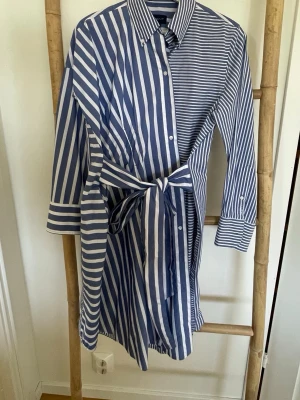 Gant Jersey Dress - Säljer en blåvit skjortklänning med olika breda ränder och knytband i midjan. Klänningen har krage, knappar framtill och långa ärmar med manschetter. Perfekt för dig som gillar en avslappnad men ändå stilren look.