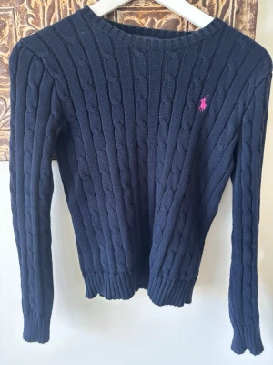 Mörkblå kabelstickad tröja Polo Ralph Lauren - Kabelstickad mörkblå tröja från Polo Ralph Lauren med rund halsringning och lång ärm. Tröjan har ett rosa broderat logotyp på bröstet och ribbade muddar vid ärmslut och nederkant. Perfekt för dig som gillar klassisk och stilren look. SKRIV OM FRÅGOR/PRISFÖRLSAG❤️❤️❤️-köpt begagnad så vet ej om äkta!
