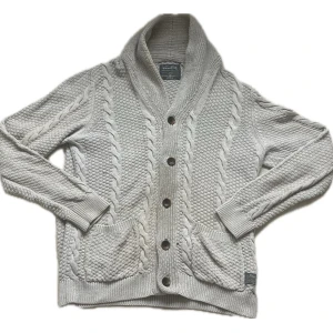 Beige Cardigan - Ljusgrå kabelstickad kofta från Vintage Gym med sjalkrage och stora bruna knappar. Koftan har två framfickor och ribbade muddar. Snygg strukturstickning och mjukt material som känns riktigt cozy.