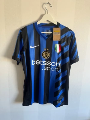 Inter Milan matchtröja Nike blå/svart - Snygg Inter Milan matchtröja från Nike i blått och svart med guldiga detaljer. Tröjan har klubbmärke, sponsorlogga och italienska mästarmärket på bröstet. Tillverkad i lätt och ventilerande material, perfekt för fotbollsträning eller match.