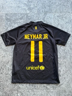 Barcelona Neymar Jr matchtröja svart - Snygg svart Barcelona matchtröja från Nike med Neymar Jr och nummer 11 i gult på ryggen. Framsidan har klubbmärke, Nike-logga och Qatar Foundation-tryck. Tillverkad i lätt och ventilerande Dri-FIT-material, perfekt för fotboll eller träning.