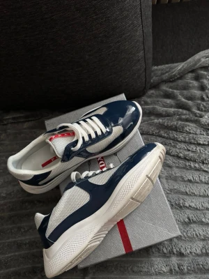 Prada sneakers i blått och vitt - Snygga sneakers från Prada i blankt marinblått och vitt med meshdetaljer och vita snören. Skorna har en chunky vit sula och röda Prada-loggor på plösen och insidan. Perfekta för dig som vill ha en clean och exklusiv look.