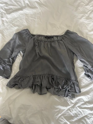 Grå offshoulder topp med volang - Säljer en grå offshoulder topp med volangdetaljer längs nederkanten och vid ärmsluten. Toppen har en loose passform och vida trekvartsärmar som ger en avslappnad vibe. Perfekt att styla med jeans eller kjol för en trendig look.