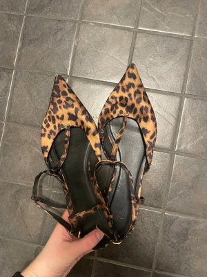 Leopardmönstrade ballerinaskor H&M - Snygga ballerinaskor från H&M med spetsig tå och leopardmönster i brunt och svart. Skorna har tunna remmar med spänne runt ankeln och är tillverkade i textilmaterial. Perfekta för dig som vill sticka ut med ett djurmönstrat statement.