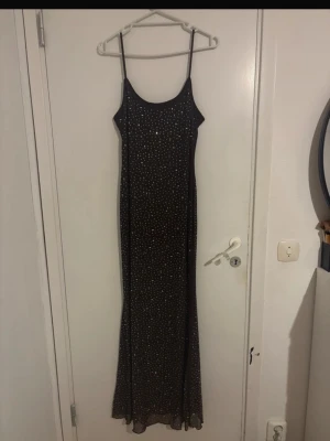 Svart långklänning med glitterdetaljer - Elegant svart långklänning med tunna axelband och glittriga paljettdetaljer över hela klänningen. Klänningen har en rak, smal passform och är perfekt för speciella tillfällen. Materialet känns lätt och följsamt.