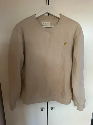 Beige sweatshirt från Pier One - Säljer en stilren beige sweatshirt från Pier One med rund halsringning och diskret gul broderad logga på bröstet. Tröjan har långa ärmar, ribbade muddar och är tillverkad i mjukt material som passar perfekt till en avslappnad look.