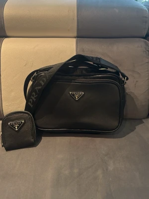 Svart axelväska från Pr$da - Säljer en svart axelväska från Pr$da med tillhörande liten plånbok. Väskan har en rektangulär form, justerbar axelrem med Prda logga och klassiskt metalltriangelmärke på både väskan och plånboken. Materialet är nylon med läderdetaljer och silverfärgade metalldelar.