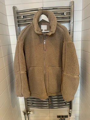 Beige teddyjacka med dragkedja - Mysig beige teddyjacka från HM  med hög krage och dragkedja framtill. Jackan har två stora fickor på framsidan och är gjord i fluffigt teddy-material som håller dig varm. Perfekt för kalla dagar och ger en avslappnad, trendig look.