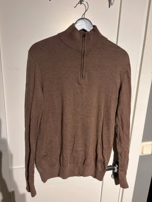 Brun halvzip-tröja Dressmann - Snygg brun långärmad tröja från Dressmann med halvzip och hög krage. Tillverkad i mjukt och tunt material, perfekt för lager på lager. Enkel och stilren design utan mönster, passar till många olika outfits.