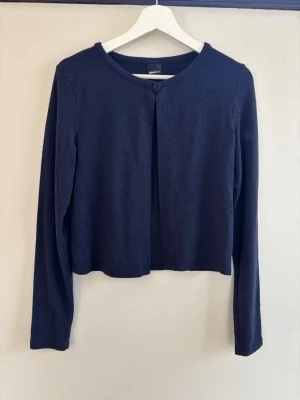 Mörkblå kofta från Gina Tricot - En enkel och stilren mörkblå kofta från Gina Tricot med rund halsringning och en knapp framtill. Koftan har långa ärmar och är tillverkad i ett mjukt material som passar perfekt till lager-på-lager. Kortare modell som ger en modern look. Passar som S/M