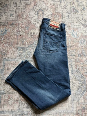 Diesel blåa raka jeans - Säljer ett par klassiska blå jeans från Diesel med raka ben och femficksmodell. Jeansen har en snygg tvättad look och tydliga sömmar på bakfickorna. Tillverkade i slitstark denim, perfekta för en avslappnad stil.