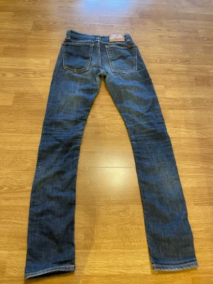Mörkblå jeans från Nudie Jeans - Säljer ett par mörkblå jeans från Nudie Jeans med klassisk femficksmodell och raka ben. Jeansen har kontrastsömmar och en läderpatch bak i midjan. Tillverkade i slitstark denim, perfekta för en avslappnad och stilren look.