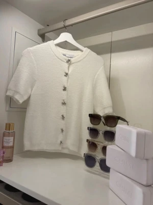Vit kortärmad kofta från Zara - Söt vit kofta från Zara med korta ärmar och dekorativa rosettknappar framtill. Koftan har en mjuk, stickad struktur och rund halsringning. Perfekt för en trendig och stilren look. Aldrig använd fick i julklapp men va tyvärr inte min still den är i stl L men sitter som S som du ser på sista bilden.