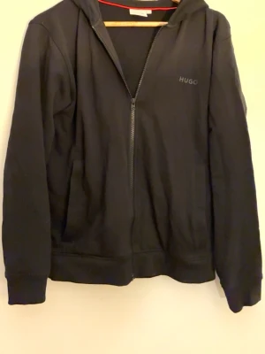 Svart hoodie med dragkedja från HUGO - Snygg svart hoodie från HUGO med dragkedja framtill och diskret logga på bröstet. Hoodien har huva, långa ärmar och två fickor framtill. Tillverkad i mjukt bomullsmaterial som känns skönt mot huden. Perfekt för en avslappnad och stilren look.