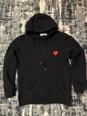 Svart hoodie Comme des Garçons Play - Svart hoodie från Comme des Garçons Play med den ikoniska röda hjärt-loggan på bröstet. Hoodien har dragsko i huvan och en klassisk känguruficka framtill. Tillverkad i mjukt bomullsmaterial som känns skönt mot huden. Perfekt för en avslappnad streetwear-look.