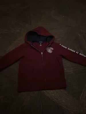Vinröd hoodie från Polo Ralph Lauren - Snygg vinröd hoodie från Polo Ralph Lauren med dragkedja framtill och vit logga på bröstet. Tryck i vitt längs ena ärmen och klassisk huva. Perfekt för en avslappnad och trendig stil.