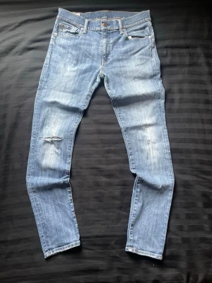 Ljusblå jeans från Polo Ralph Lauren - Säljer ett par ljusblå jeans från Polo Ralph Lauren med slitningar och ett hål på vänster knä. Jeansen har klassisk femficksmodell, raka ben och är tillverkade i bomull. Perfekta för en avslappnad och trendig look.