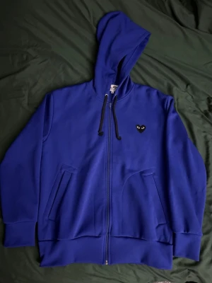 Blå Comme des Garçons Play hoodie - Snygg blå hoodie med dragkedja från Comme des Garçons Play i storlek L men passar även jätte bra i M. Har svart hjärta med ögon på bröstet och stort hjärta med ögon på ryggen. Utrustad med huva och svarta dragsnören samt två fickor framtill. Tillverkad i mjukt bomullsmaterial.