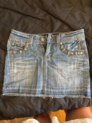 Blå jeanskjol med nitar - Snygg blå jeanskjol med slitna detaljer och rå kant nertill. Kjolen har nitar på fickorna fram och en bakficka med dekorativ söm. Klassisk femficksmodell och bälteshällor. Perfekt för en cool och avslappnad stil.