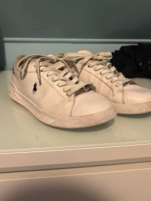 Vita låga sneakers från Polo Ralph Lauren - Säljer ett par klassiska vita sneakers från Polo Ralph Lauren med den ikoniska mörkblåa loggan på sidan. Skorna har vita snören, låg profil och är tillverkade i slätt läder. Perfekta för en clean och stilren look.