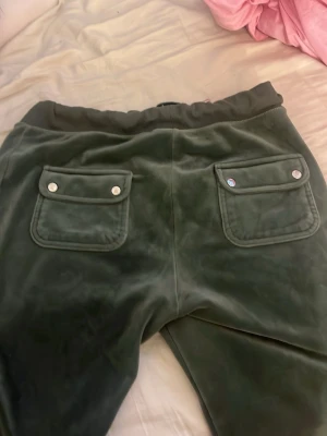 Gröna  Juicy Couture - Mörkgröna mjukisbyxor i velour från Juicy Couture. Byxorna har bred resår i midjan och två bakfickor med lock och silvriga knappar. 