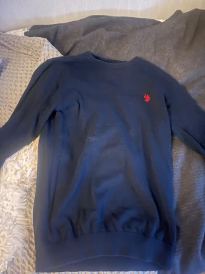Mörkblå tröja från Polo Assn - Snygg mörkblå tröja från Polo Assn med rund halsringning och röd broderad logga på bröstet. Tröjan har långa ärmar och ribbade muddar vid ärmslut och nederkant. Perfekt för en stilren och enkel look.