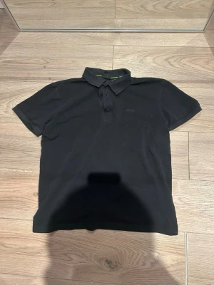 Svart pikétröja från BOSS - Klassisk svart pikétröja från BOSS med korta ärmar och krage. Tröjan har knäppning framtill och diskret BOSS-logga på bröstet. Tillverkad i mjukt bomullsmaterial och har en stilren look med gröna detaljer på insidan av kragen.