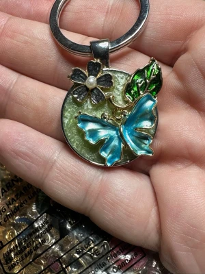 Nyckelring med fjäril och blomma - Unik nyckelring med rund metallbricka dekorerad med en blå fjäril i emalj, svart blomma med pärldetalj och grön bladdekor. Färgerna är metallic och detaljerna är i guldton. Perfekt accessoar för att piffa upp nyckelknippan.