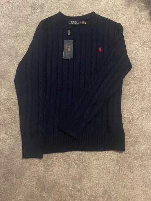 Mörkblå kabelstickad tröja Polo Ralph Lauren - Mörkblå kabelstickad tröja från Polo Ralph Lauren med rund halsringning och röd broderad logga på bröstet. Tröjan har långa ärmar och ribbade muddar vid ärmslut och nederkant. Klassisk och stilren design i mjukt material.