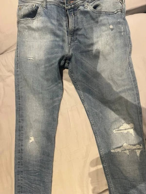 Ljusblå slitna replay jeans - Säljer ett par ljusblå jeans med slitningar och hål på benen. Jeansen har fem fickor, normal midja och smal passform. Snygga detaljer med slitna partier framtill och klassiska bakfickor med broderade sömmar. Perfekta för en avslappnad streetstil.