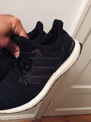 Svarta Adidas Ultra Boost sneakers - Säljer ett par svarta Adidas Ultra Boost sneakers med stickad ovandel och vita Boost-sulor. Skorna har klassiska tre ränder på sidan, svart plastdetalj och svart yttersula. Perfekta för dig som gillar stilrena och bekväma sneakers.