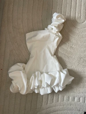 Vit axelbandslös kortklänning - Trendig vit axelbandslös kortklänning från Fashion Nova med volangdetaljer i nederkant och en stor rosett på ena axeln. Klänningen har en figurnära passform och är perfekt för dig som vill sticka ut. Tillverkad i ett stretchigt material som formar sig efter kroppen. ”wilma ruffle mini dress white”. Oanvänd med prislappen kvar!! Perfekt till studenten. Priset kan diskuteras 