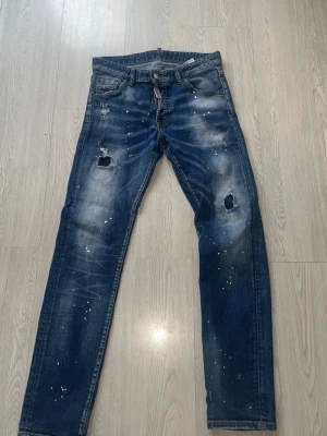 Mörkblå distressed jeans med färgstänk - Säljer ett par mörkblå jeans med slitningar och hål på knäna samt coola färgstänk över hela byxorna. Jeansen har fem fickor och raka ben. Perfekta för dig som gillar en avslappnad och trendig streetstil.