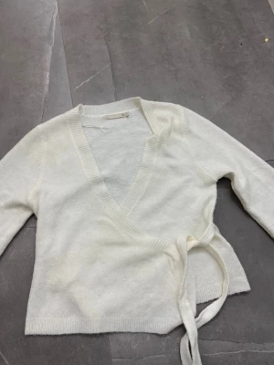 Vit omlottkofta från lager 157  - Säljer en mjuk och stilren vit stickad omlottkofta från lager 157  Koftan har långa ärmar, ribbade muddar och knytskärp i midjan för en snygg passform. Perfekt att slänga på sig över en topp för en enkel men trendig look.