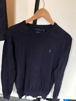 Mörkblå tröja från Polo Ralph Lauren - Klassisk mörkblå tröja från Polo Ralph Lauren med rund halsringning och diskret broderad logga på bröstet. Tillverkad i mjukt material och har ribbade muddar vid ärmslut och nederkant. Perfekt för en stilren och enkel look.
